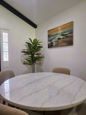 A round marble table with four chairs, a potted plant, a painting of a beach scene, and a white door with a window. - Lake Lanier Getaway. Hot Tub & Fire Pit. 5-Min Walk to Lake Access (Flowery Branch)