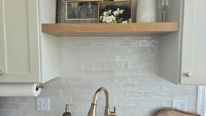 Eine Küche mit weiß gefliester Spritzschutzwand, einem Holzbodenregal mit gerahmten Bildern, einem Messing-Wasserhahn und einer Holz-Schneidebrett.