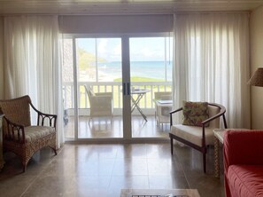 Ein Zimmer mit Meerblick, eine Glastür, Korbstühle, ein rotes Sofa und ein kleiner Tisch.