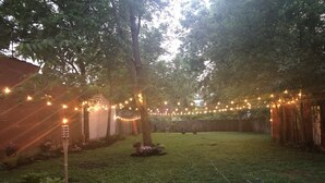 Un patio trasero con terraza de madera, luces colgantes y zona ajardinada.