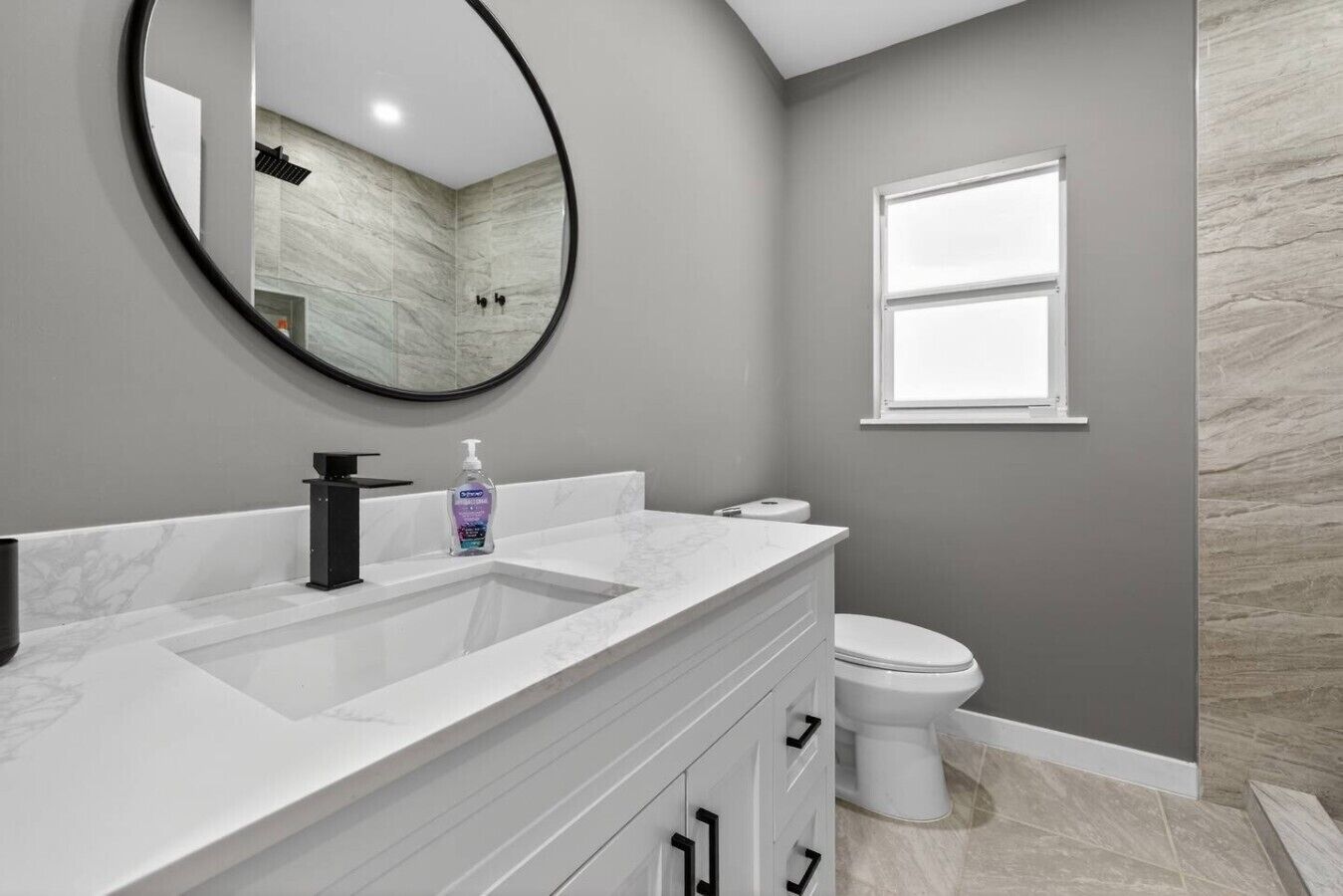 A bathroom with a white vanity, a round mirror, a toilet, and a window.