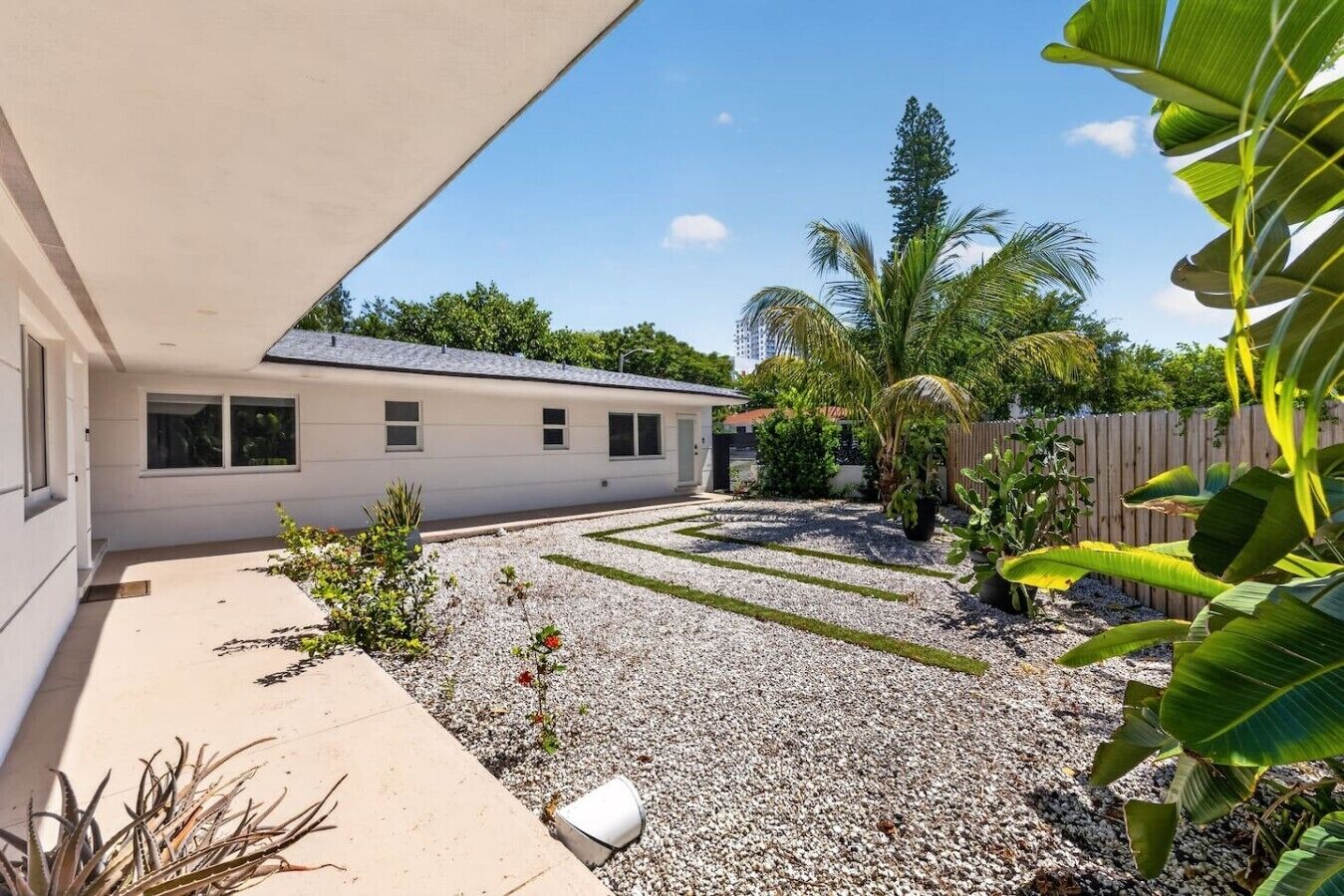 Una propiedad residencial con un patio cubierto, un camino de entrada de grava y diversas plantas tropicales.