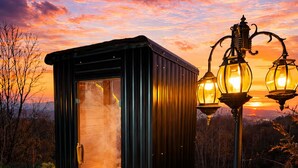 A small, dark-colored outdoor structure with a door, a bench, and a stone pathway with scattered rose petals. - Cozy Chalet with Jacuzzi, Heated pool & Sauna Montebliss (Miljevići)