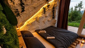 A bedroom with a stone wall, a bed with dark bedding, a bedside table with a lamp, and a small plant. - Cozy Chalet with Jacuzzi, Heated pool & Sauna Montebliss (Miljevići)