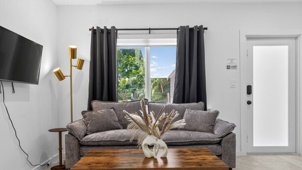 Ein Wohnzimmer mit einem grauen Sofa, einem Holztisch, einer Stehlampe und einem an der Wand befestigten Fernseher.
