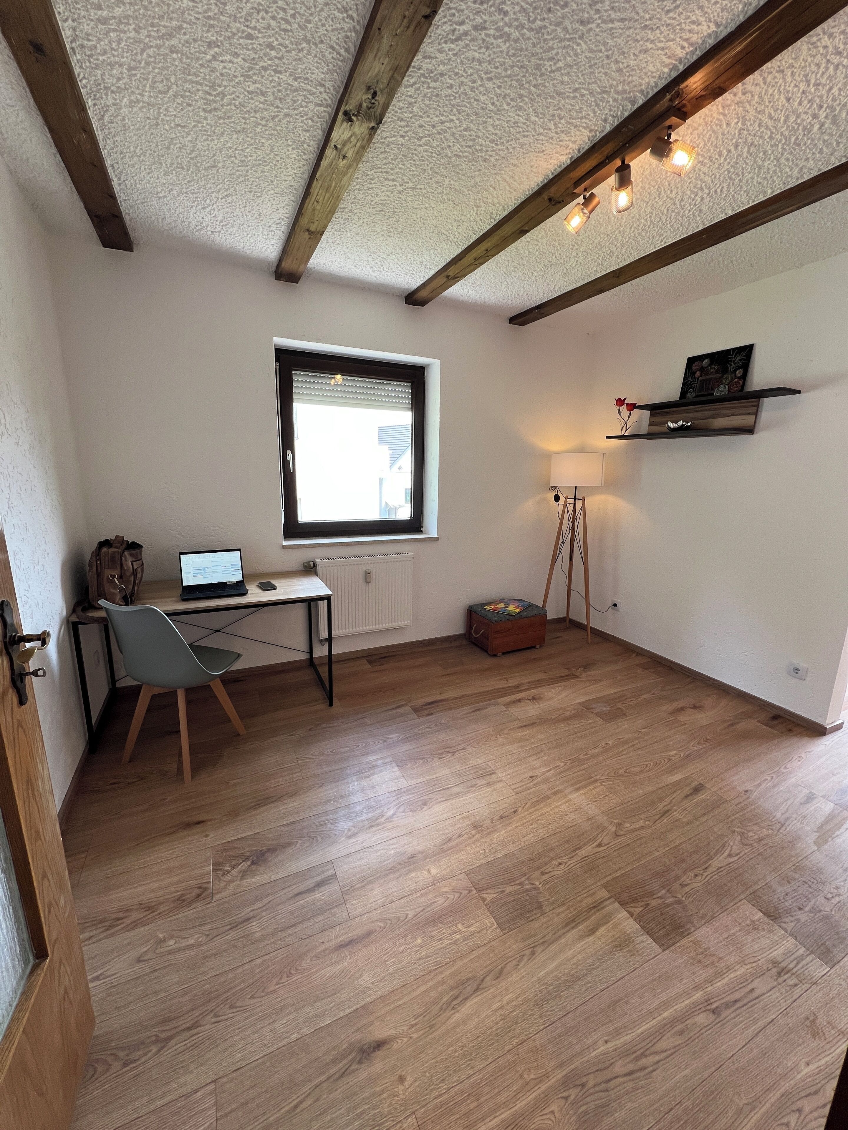 A room with wooden flooring, a desk with a computer, a chair, a radiator, a window with blinds, a shelf with decorative items, and a standing lamp.