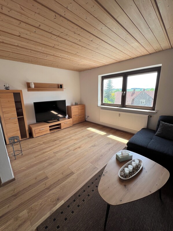 Ein modernes Wohnzimmer mit Holzboden, einem Fernseher, einem Sofa, einem Couchtisch mit Tablett und einem Fenster mit Blick auf Häuser.