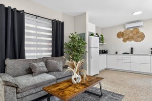 A modern living room with a grey sofa, a wooden coffee table, and a kitchen area with white cabinets and a white refrigerator. - 15 min to Beach 2 Queen Beds Free Parking U3 (Miami)
