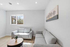 A modern living room with a grey sofa, a round coffee table, and a window with a view of trees and a building. - Beautiful 3BR Getaway in Trendy NW Calgary (Calgary)