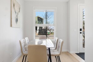 A dining area with a table and chairs, a window with a view of a building and trees, and a door with a doormat. - Stylish Family Home • 3BR + 2.5 Bath • NW Calgary (Calgary)