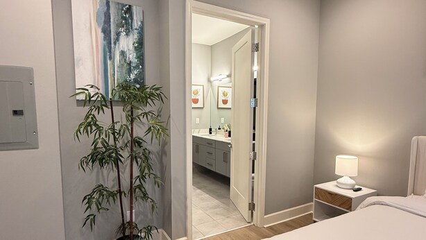 A bedroom with a bed, bedside table with a lamp, a potted plant, a mirror, and a bathroom visible through an open door. - Luxury Modern King Bed Heart of Downtown Dallas (Dallas)