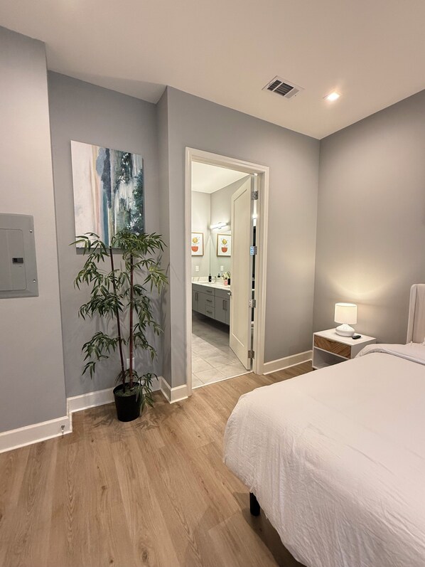 A bedroom with a bed, bedside table with a lamp, a potted plant, a mirror, and a bathroom visible through an open door.