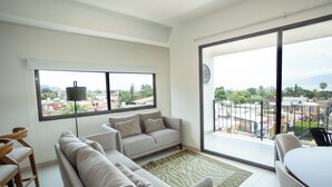 A modern living room with a sofa, a dining table, and a large window offering a city view. - Suites Santa Isabella (Ajijic)
