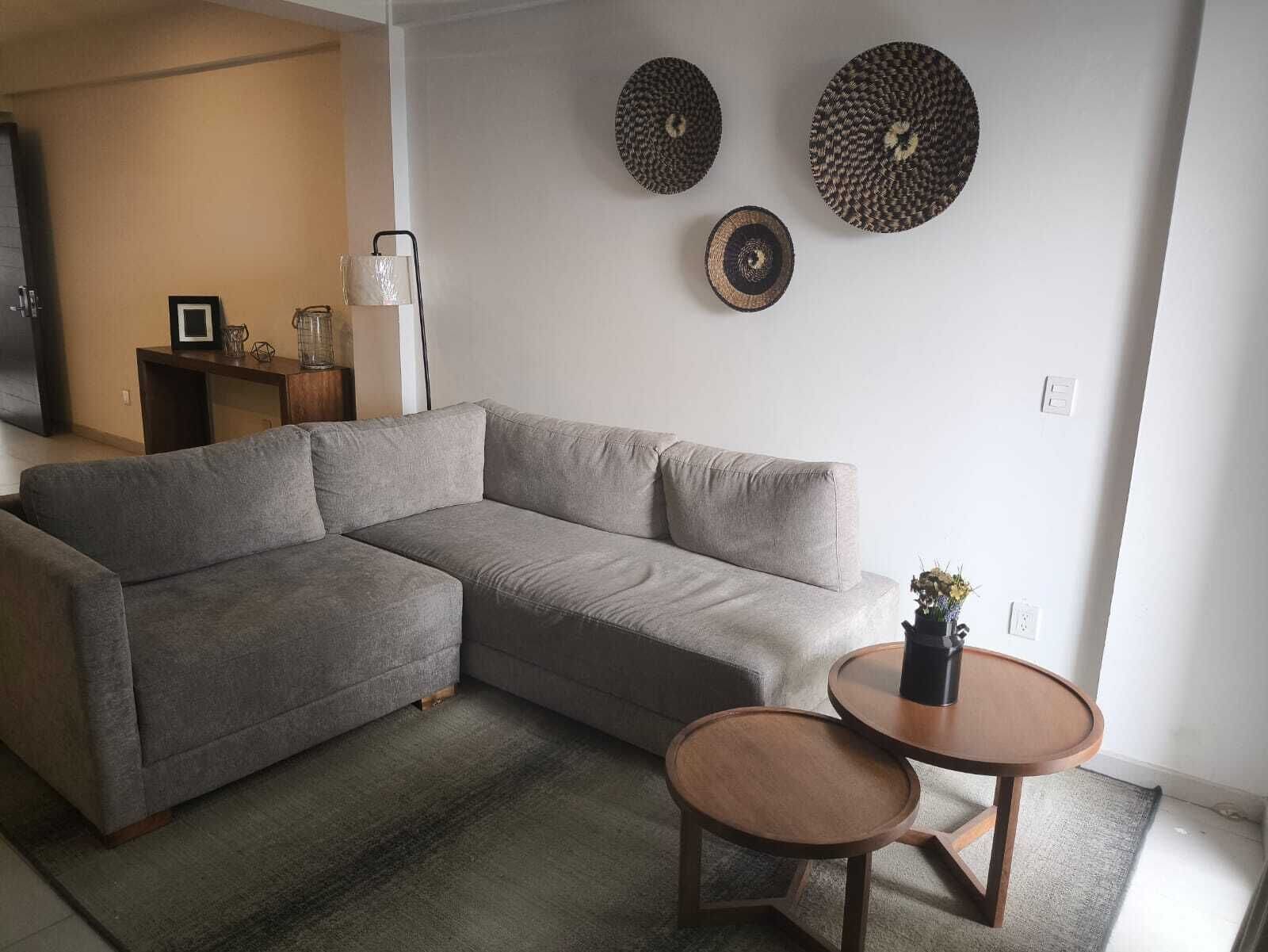Una sala de estar con un sofá seccional gris, una mesa de centro redonda de madera y dos placas decorativas colgadas en la pared.