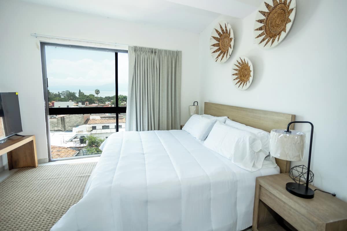 Une chambre dâhĂŽtel avec un grand lit, un tĂ©lĂ©viseur posĂ© sur un meuble en bois, une table de chevet avec une lampe, et une vue sur la ville par une grande fenĂȘtre.
