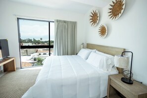 A hotel room with a large bed, a TV on a wooden stand, a nightstand with a lamp, and a view of the city through a large window. - Suites Santa Isabella (Ajijic)