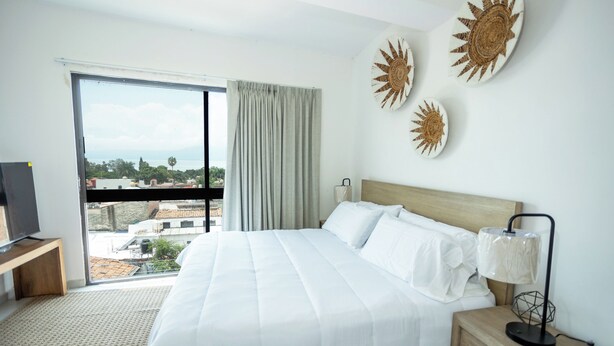 A hotel room with a large bed, a TV on a wooden stand, a nightstand with a lamp, and a view of the city through a large window. - Suites Santa Isabella (Ajijic)