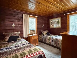 Una cabaña con dos camas, paredes de madera, una ventana con cortinas, una mesita de noche y un cuadro enmarcado.