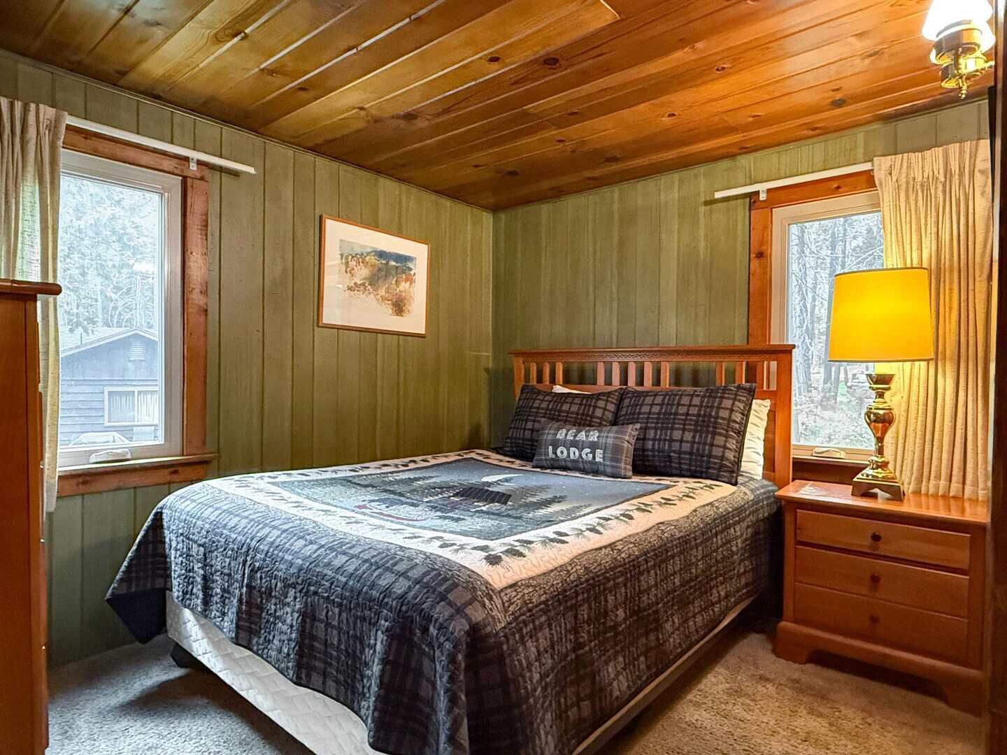 A bedroom with a wooden bed, a nightstand, a lamp, a framed picture on the wall, and a window with curtains.
