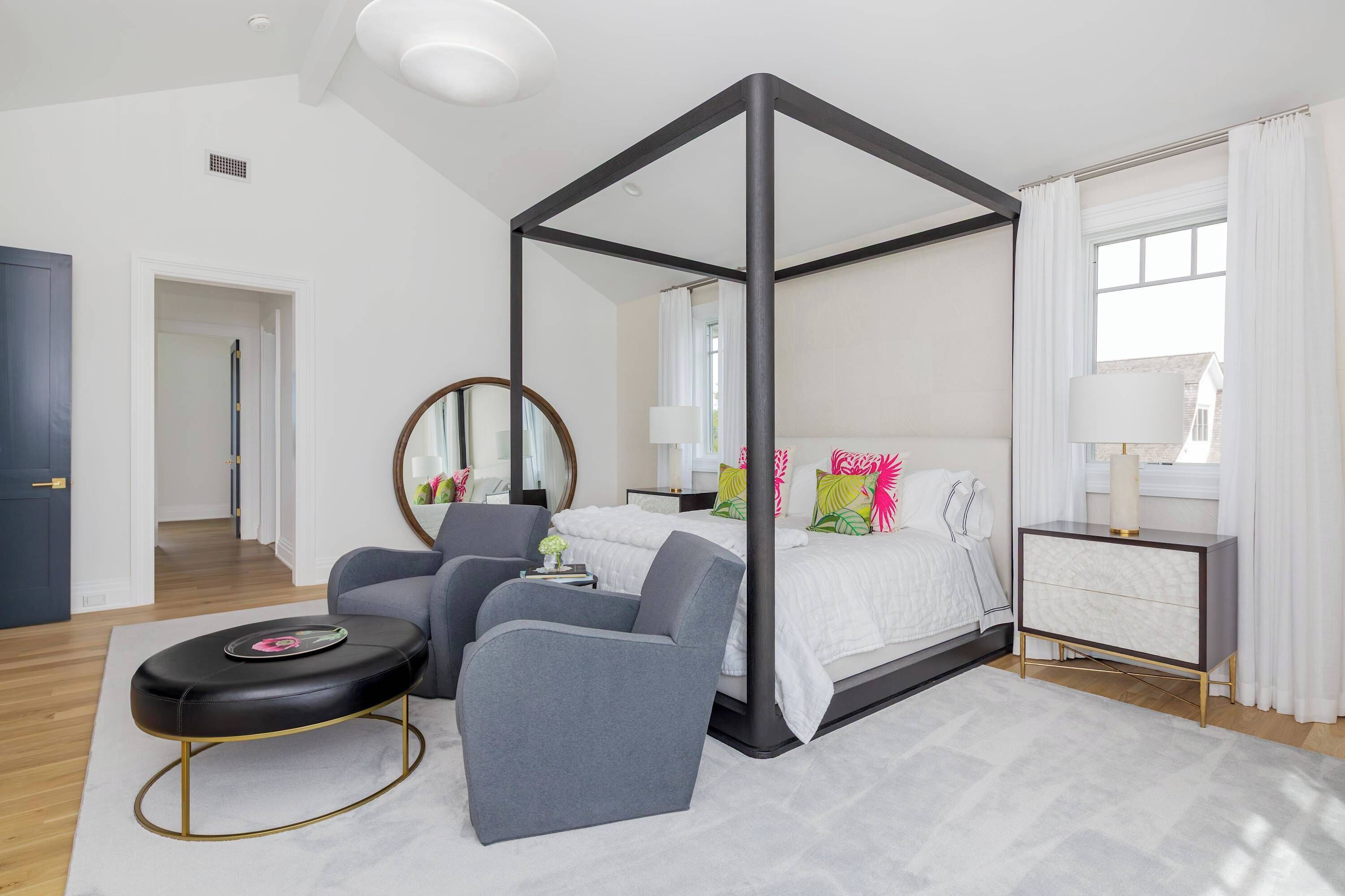 A bedroom with a four-poster bed, a bedside table, a chair, a round ottoman, and a mirror.