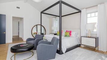 A bedroom with a four-poster bed, a bedside table, a chair, a round ottoman, and a mirror.