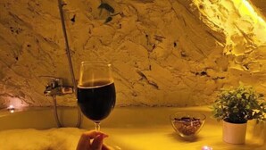 Una persona relaxant-se en una banyera, amb un got de vi, envoltada de plantes i un bol de llaminadures.