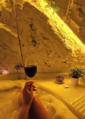 Une personne se dĂ©tend dans un bain, un verre de vin Ă la main, entourĂ©e de plantes et dâun bol de collations.