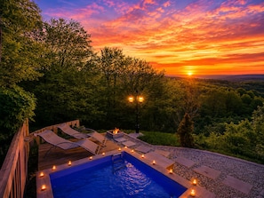 A pool area with lounge chairs and candles, surrounded by trees and a sunset view.