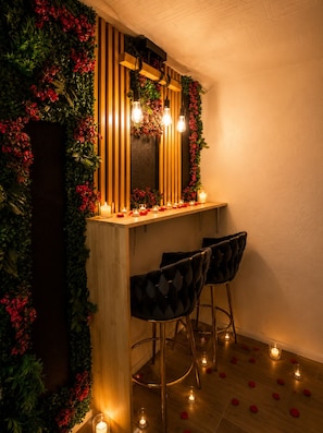 A bar area with a wooden counter, two upholstered bar stools, a mirror, and a wall adorned with greenery and red flowers.