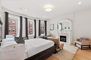 A modern bedroom with a large bed, a fireplace, a sofa, and a view of a red brick building.