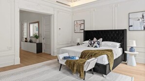 A bedroom with a large bed, a side table, a chair, a mirror, and a door leading to another room.