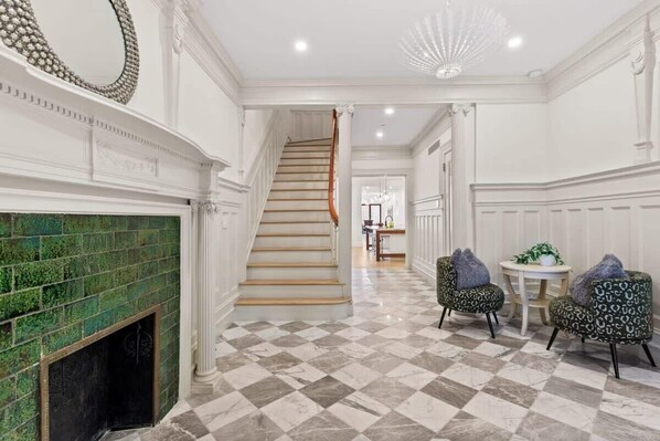 A hallway with a checkered floor, a staircase, a fireplace with green tiles, and patterned armchairs.