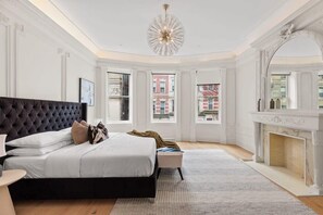 A bedroom with a large bed, a fireplace, and a view of buildings through the windows.