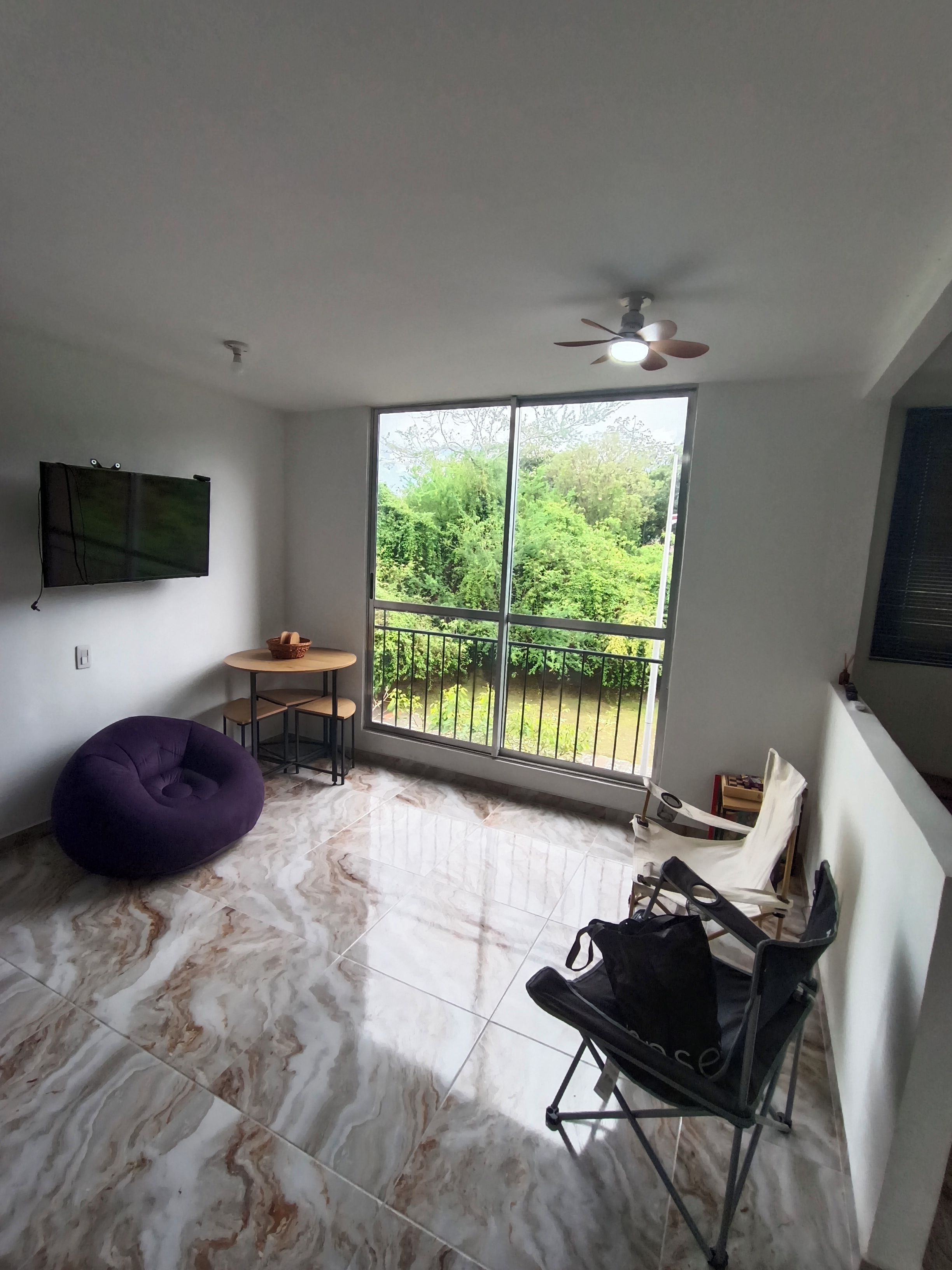 A room with a large window, a ceiling fan, a flat-screen TV, a bean bag chair, a folding chair, and a small table.
