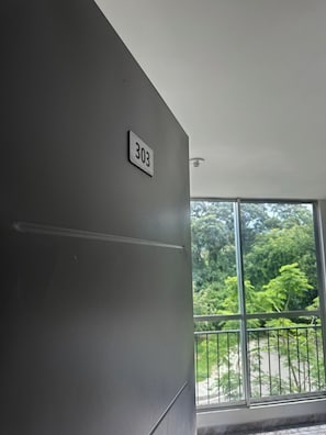 Ein Zimmer mit der Nummer '303' an der Wand, einem Fenster mit Blick auf Grünflächen und einem Balkon mit Metallgeländer.