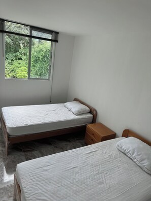 Ein Schlafzimmer mit Bett, Nachttisch, Fenster mit Jalousien und Blick auf Grünfläche.