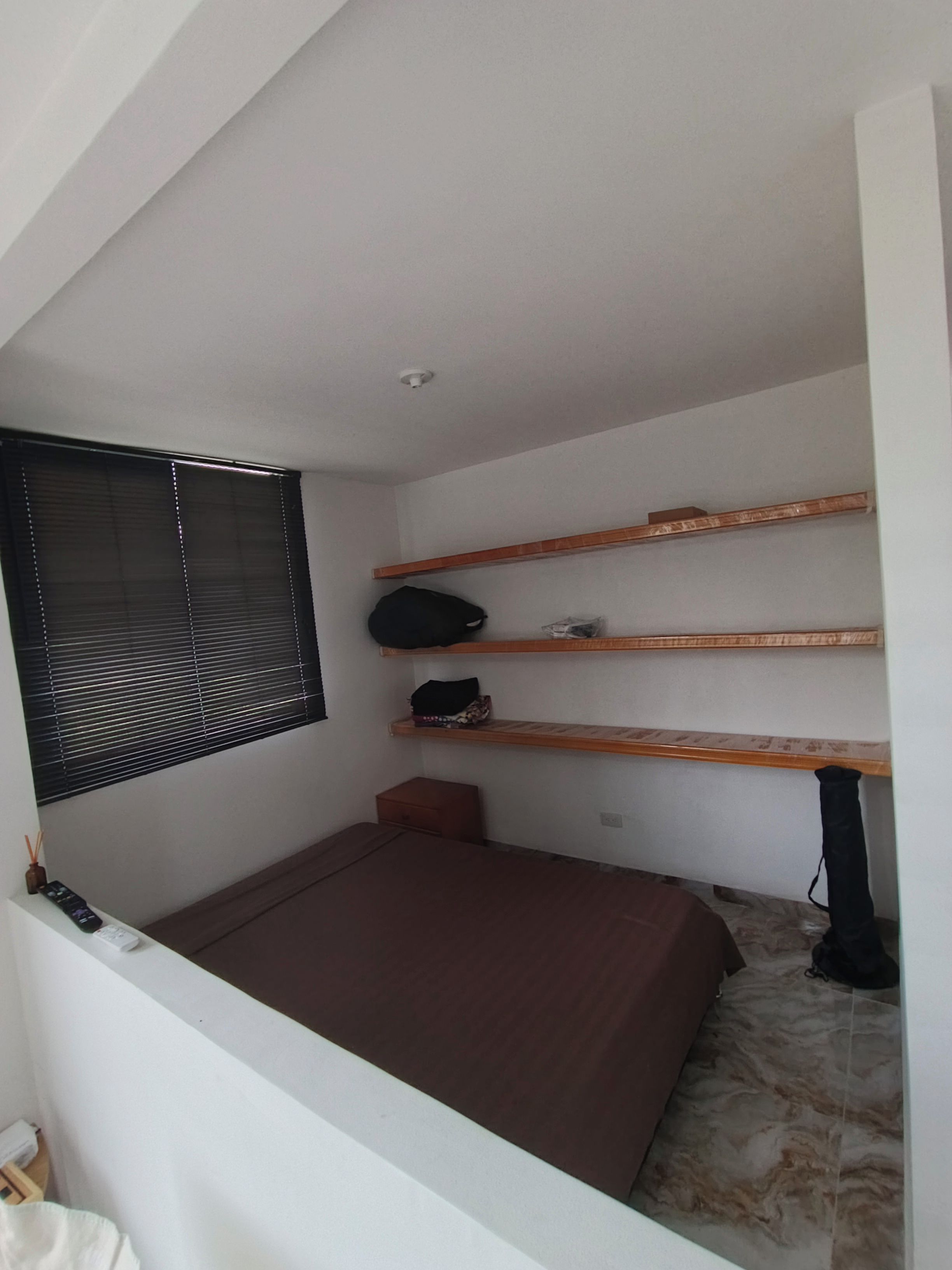 Een kleine, compacte kamer met een enkel bed, een raam met zonwering en ingebouwde planken.