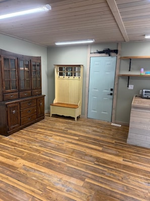 Una habitación con piso de madera, un mueble de madera grande, un banco, una puerta y un estante con objetos.