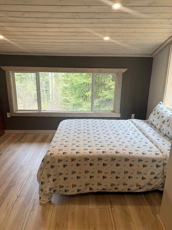 Un dormitorio con un ventanal grande, suelo de madera y una cama con ropa de cama estampada.