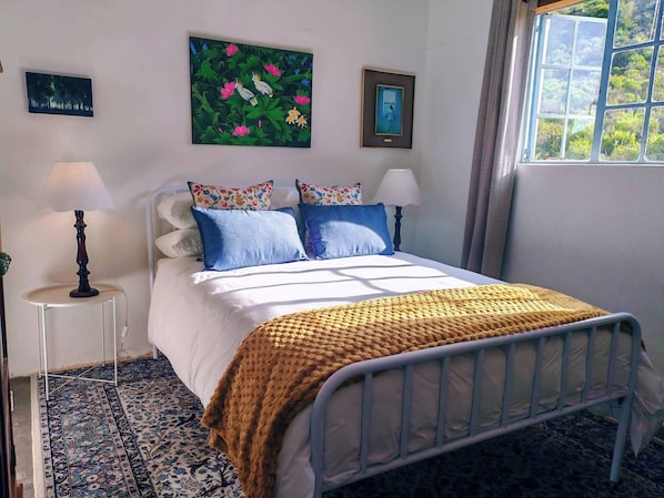 A bed with white bedding, two pillows, a knitted throw, and a metal frame. There are two wall-mounted lamps, a framed painting, and a window with a view of greenery.