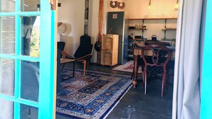Une pièce dotée d’une grande fenêtre, d’une chaise en bois, d’un tapis, d’un meuble en bois et d’un coin cuisine avec des étagères et une bouilloire.