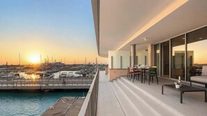 Un balcón con vistas a un puerto deportivo al atardecer, un área de comedor con sillas y mesas, y un diseño arquitectónico moderno.