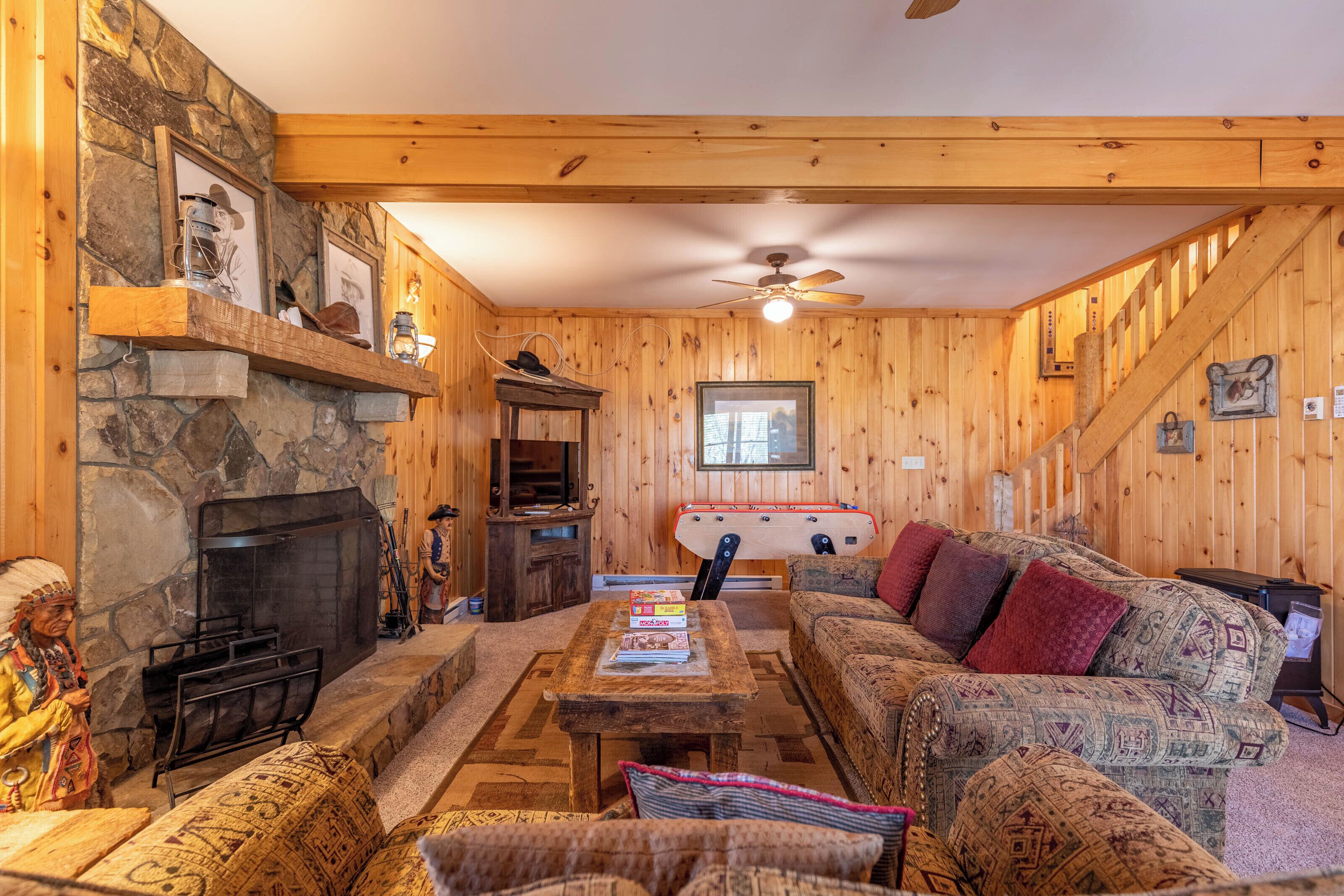 A cozy living room with a stone fireplace, wooden walls, a ceiling fan, and a staircase.