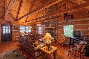 Interior de una cabaña de troncos con chimenea, muebles de madera y un televisor.