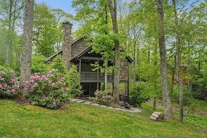 A rustic cabin with a stone chimney, a balcony, and surrounded by lush greenery.