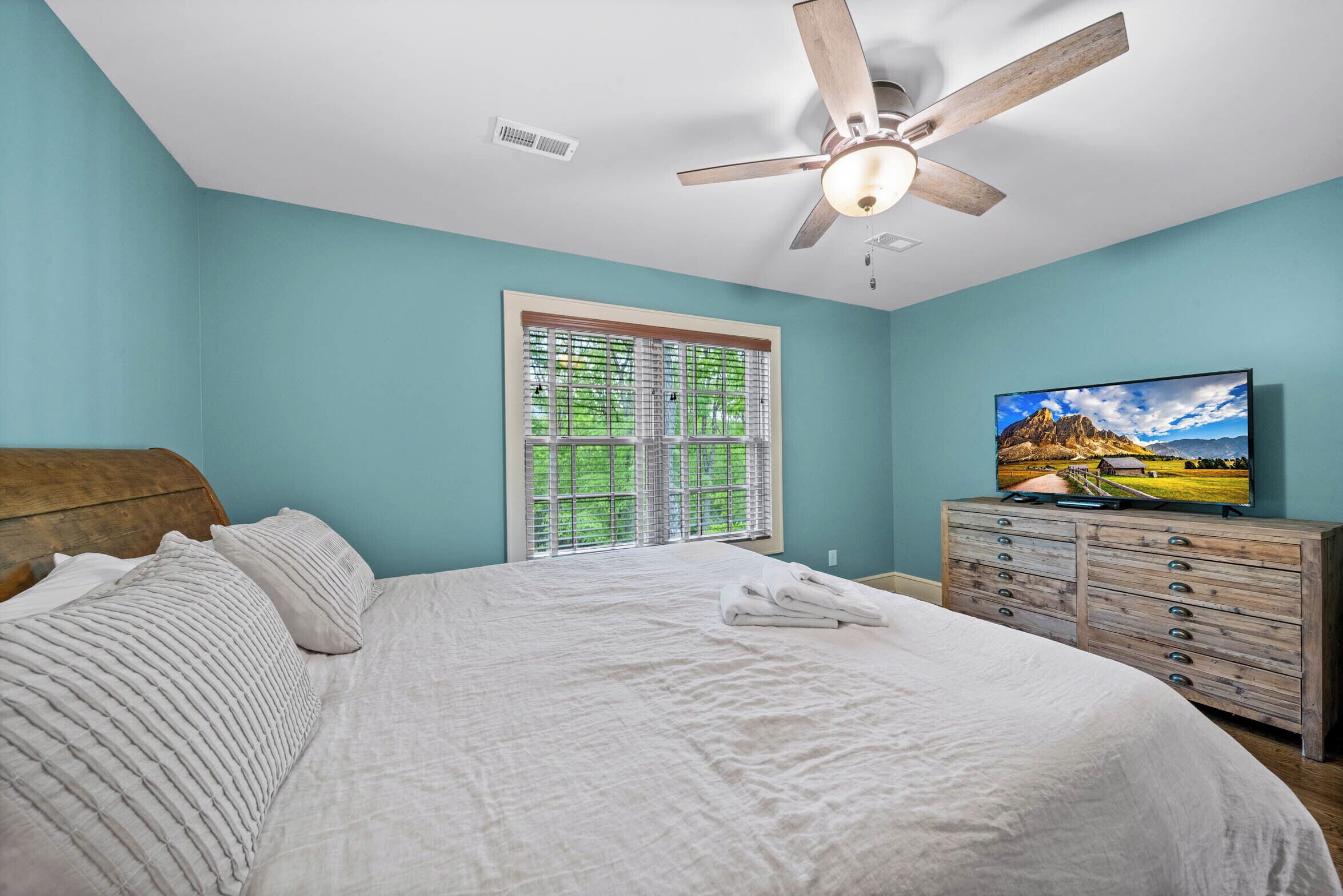 Une chambre à coucher avec un lit, un ventilateur de plafond, un téléviseur et une fenêtre avec des stores.
