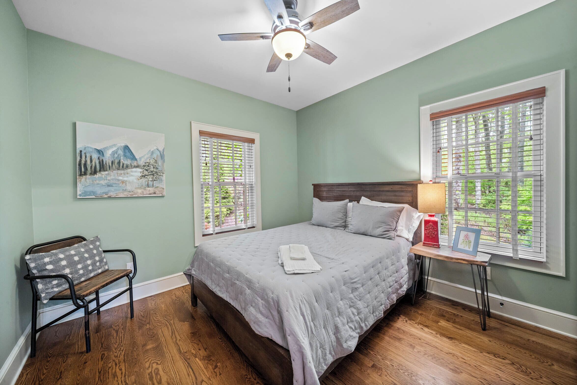 A bedroom with a bed, a bench, a ceiling fan, and two windows with blinds.