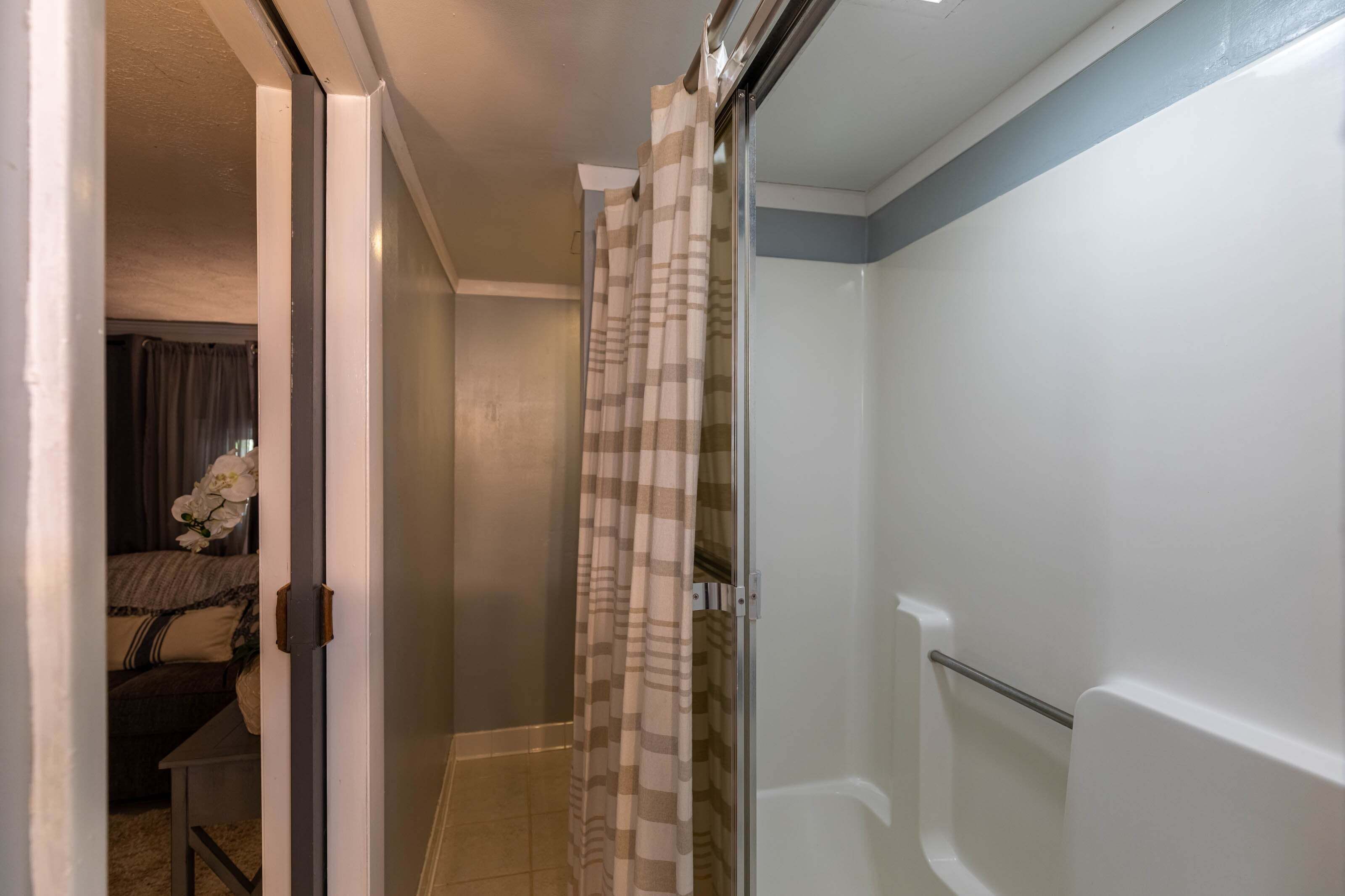 A bathroom with a shower, a toilet, and a sink. There is a door leading to another room with a bed and a window.