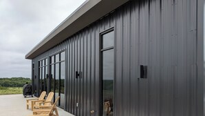 Un edificio moderno con grandes ventanales, una zona de estar al aire libre techada con sillas de madera y vistas al paisaje.