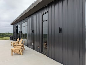 A modern building with large windows, a covered outdoor seating area with wooden chairs, and a view of the landscape.